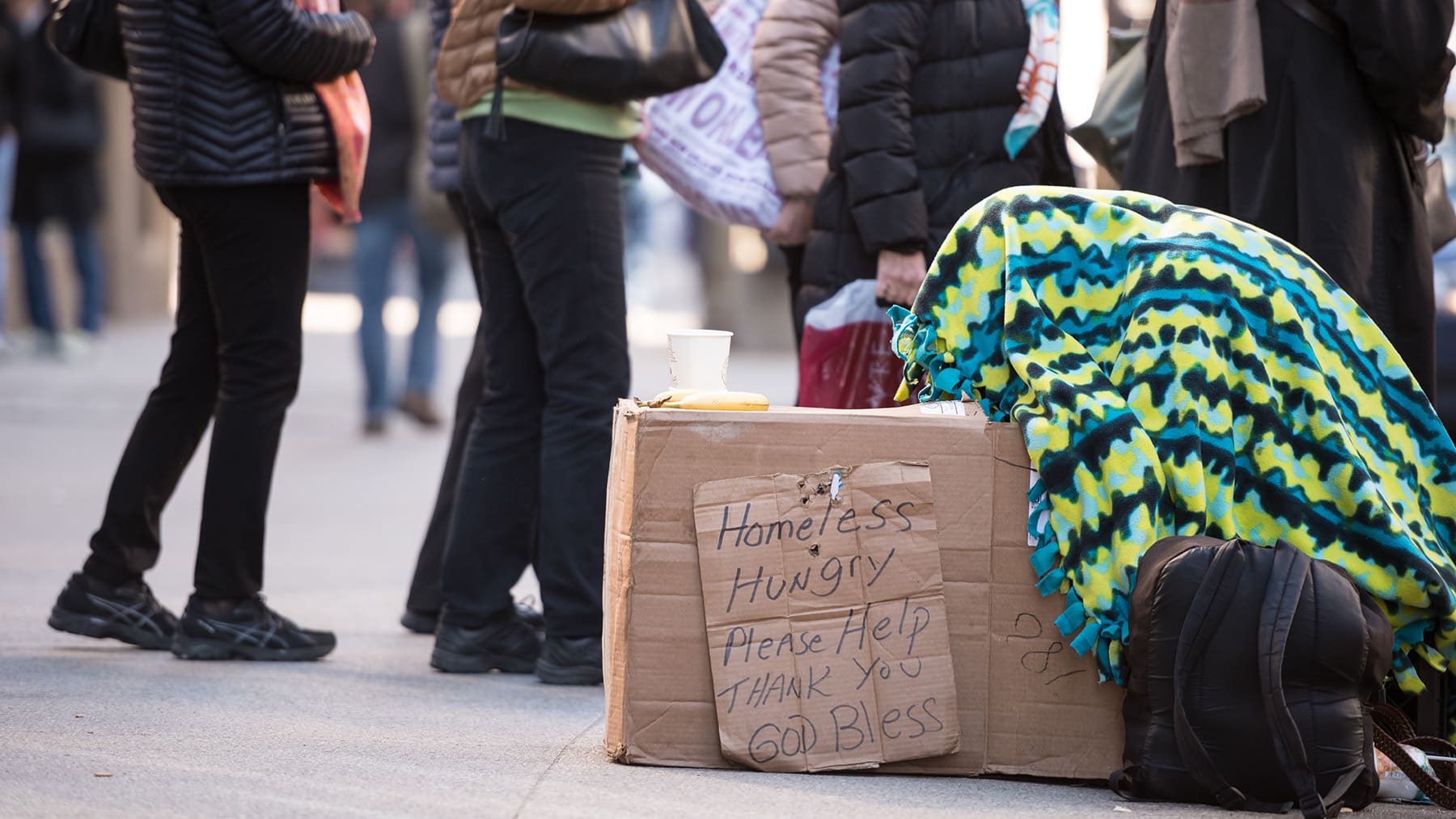 Person under a blanket with a sign asking for help as people walk past.