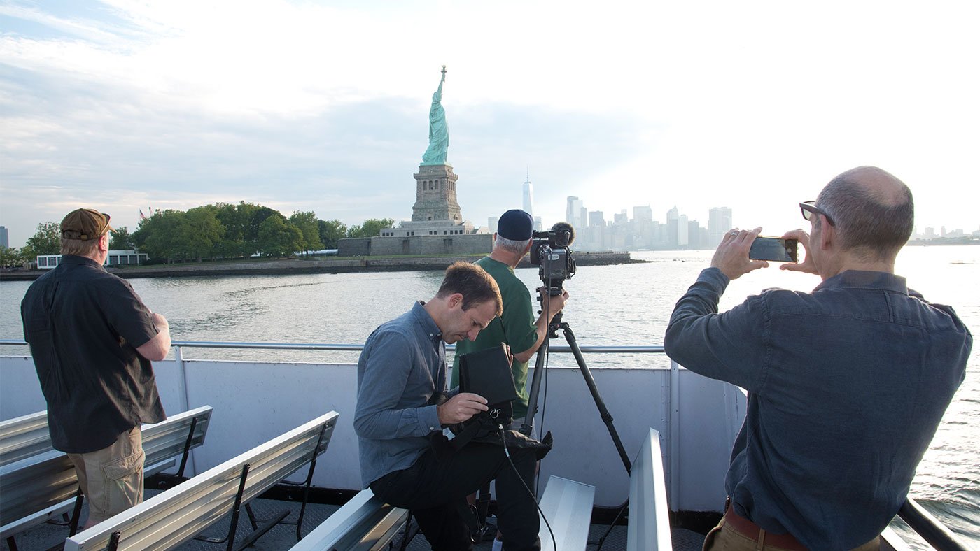 Director of Photography Tim Boyd captures a sunrise over Manhattan aboard a ferry bound for Liberty Island while Executive Producer Dan Protess watches through a viewfinder and Geoffrey Baer takes a photo