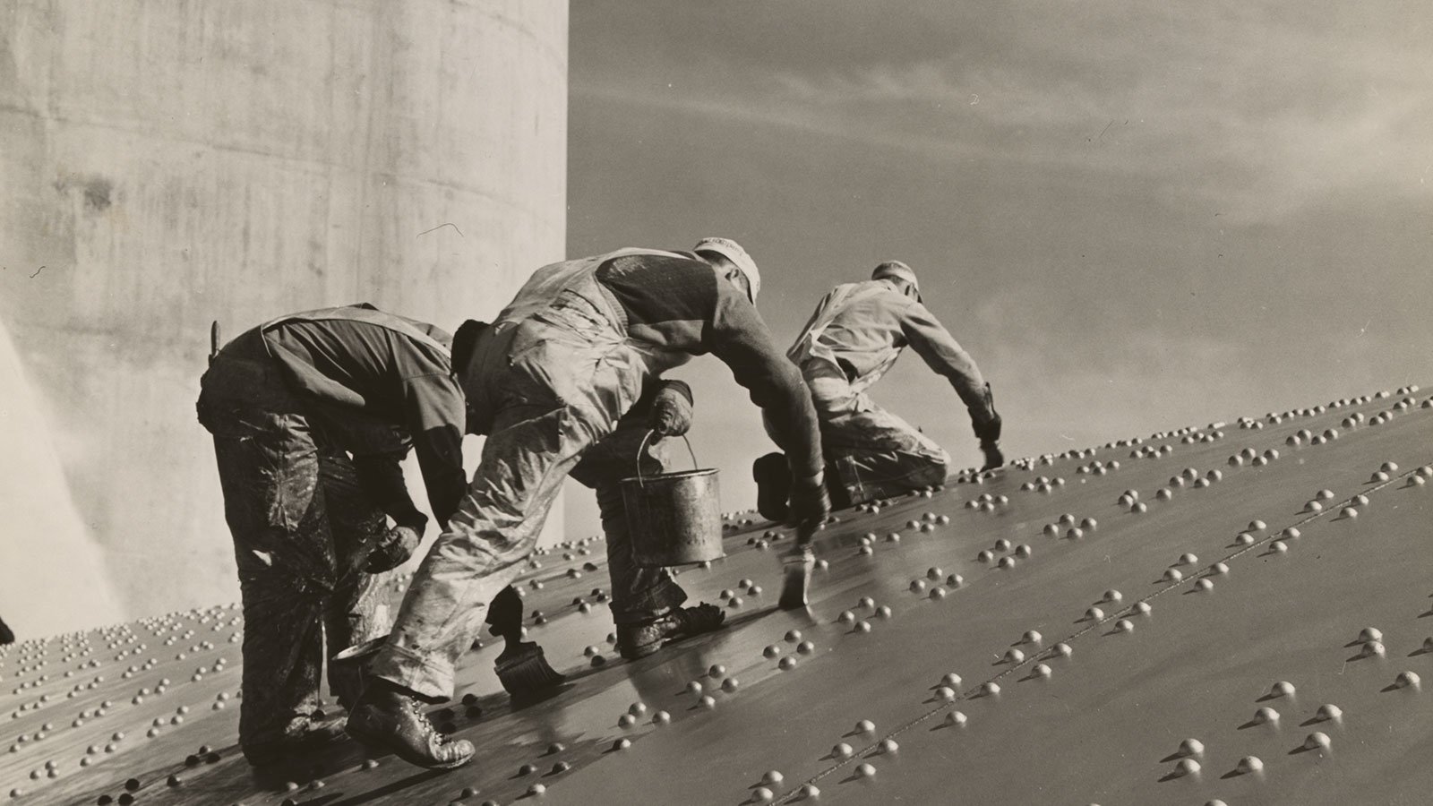 Three construction workers putting a coat of paint on a slanted wall of riveted-steel plates on the Hoover Dam spillway