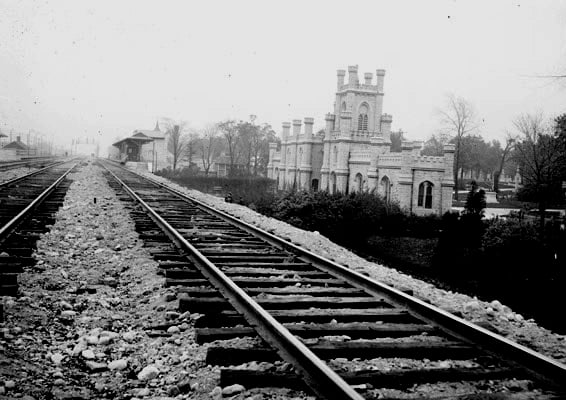 Train tracks run past a Gothic structure towards a station in a black and white photo