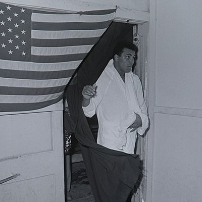 Muhammad Ali coming through doorway draped in a United States flag. Fifth Street Gym. Miami, FL. February 25, 1971. Photo: Courtesy of Charles Trainor