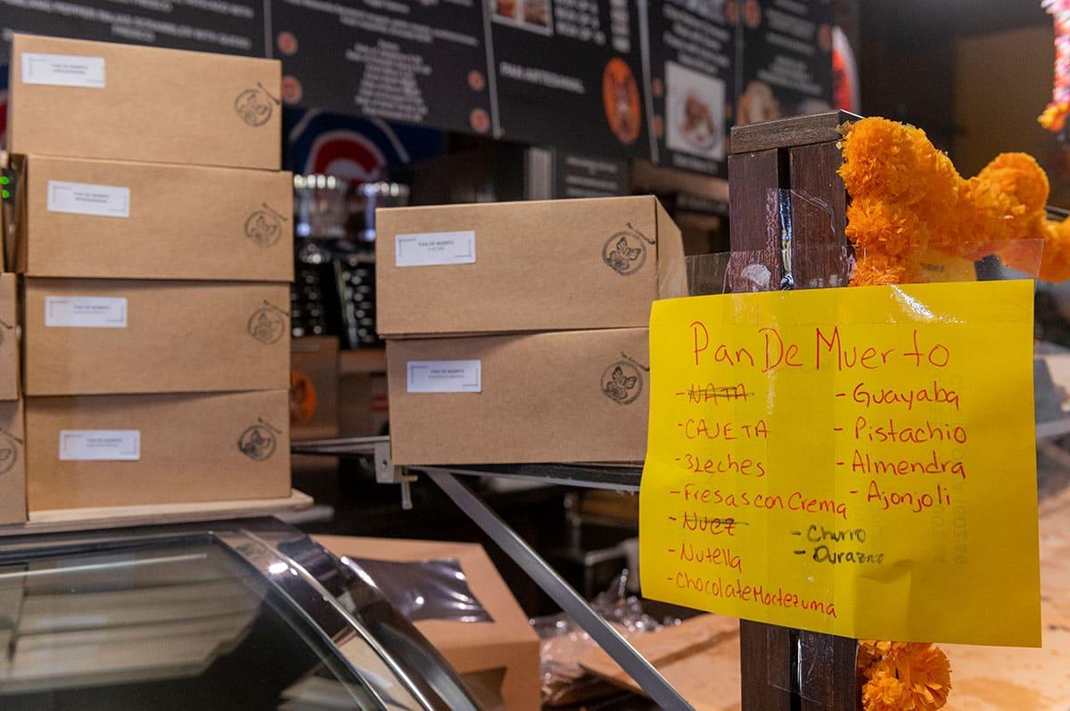A handwritten sign showing the available flavors of pan de muerto, with boxes stacked behind