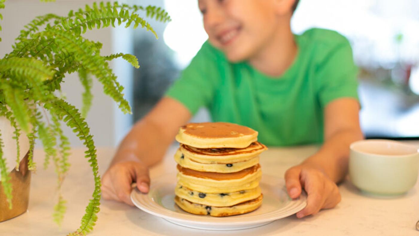 A kid smiles while grabbing a plate with a stack of pancakes