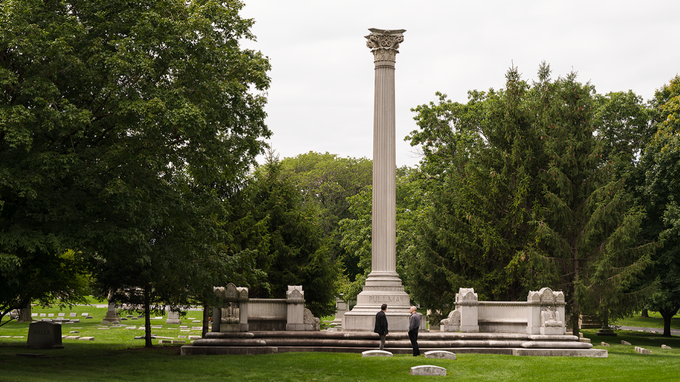 Two men stand in front of a column that is part of a memorial in a cemetery