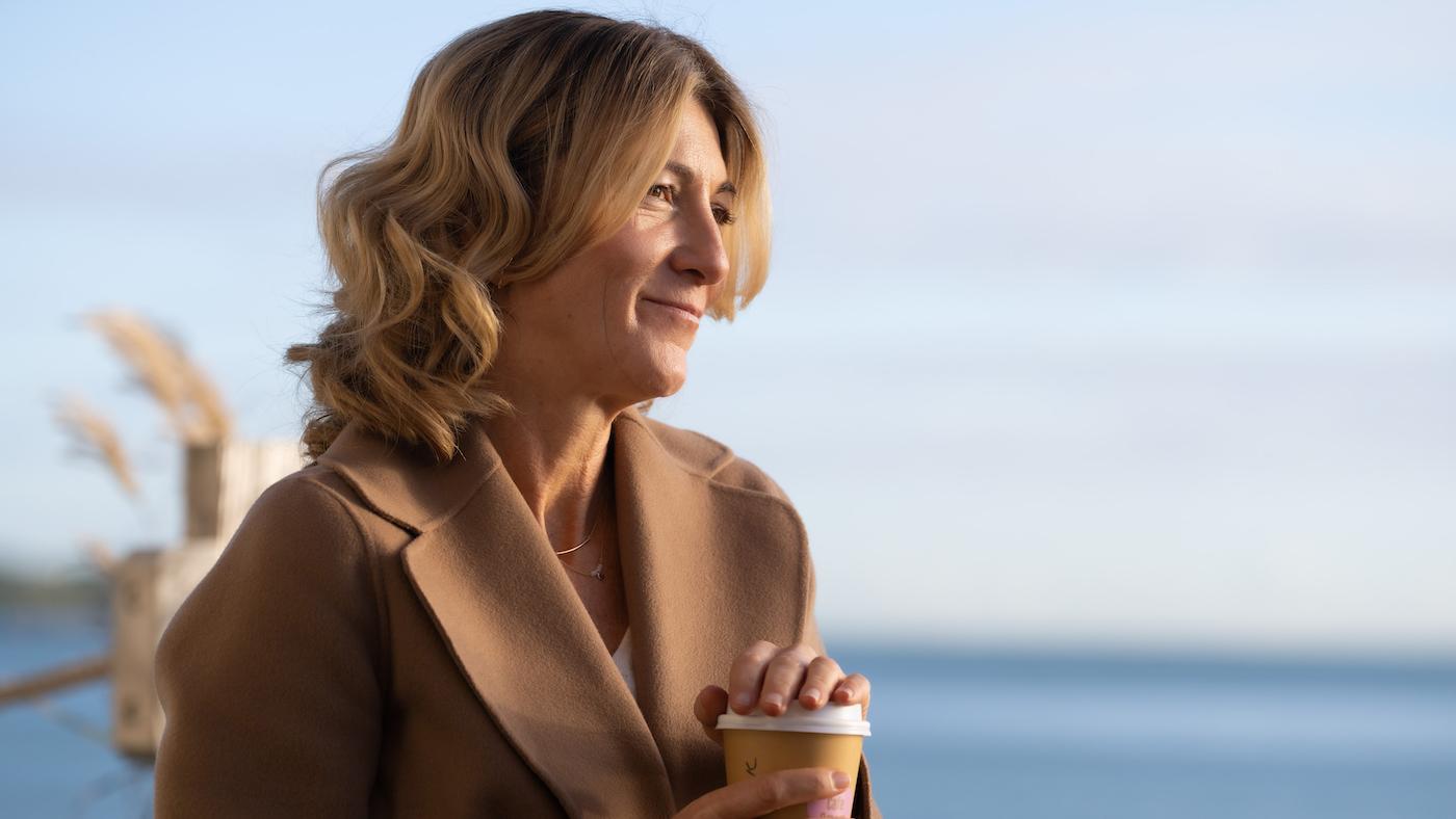 Rosaline holds a cup of coffee and smiles in front of the ocean