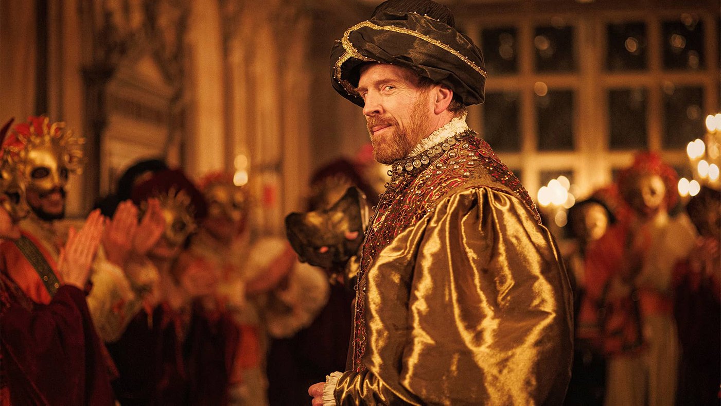 King Henry VIII smirks in front of a crowd of masked revelers