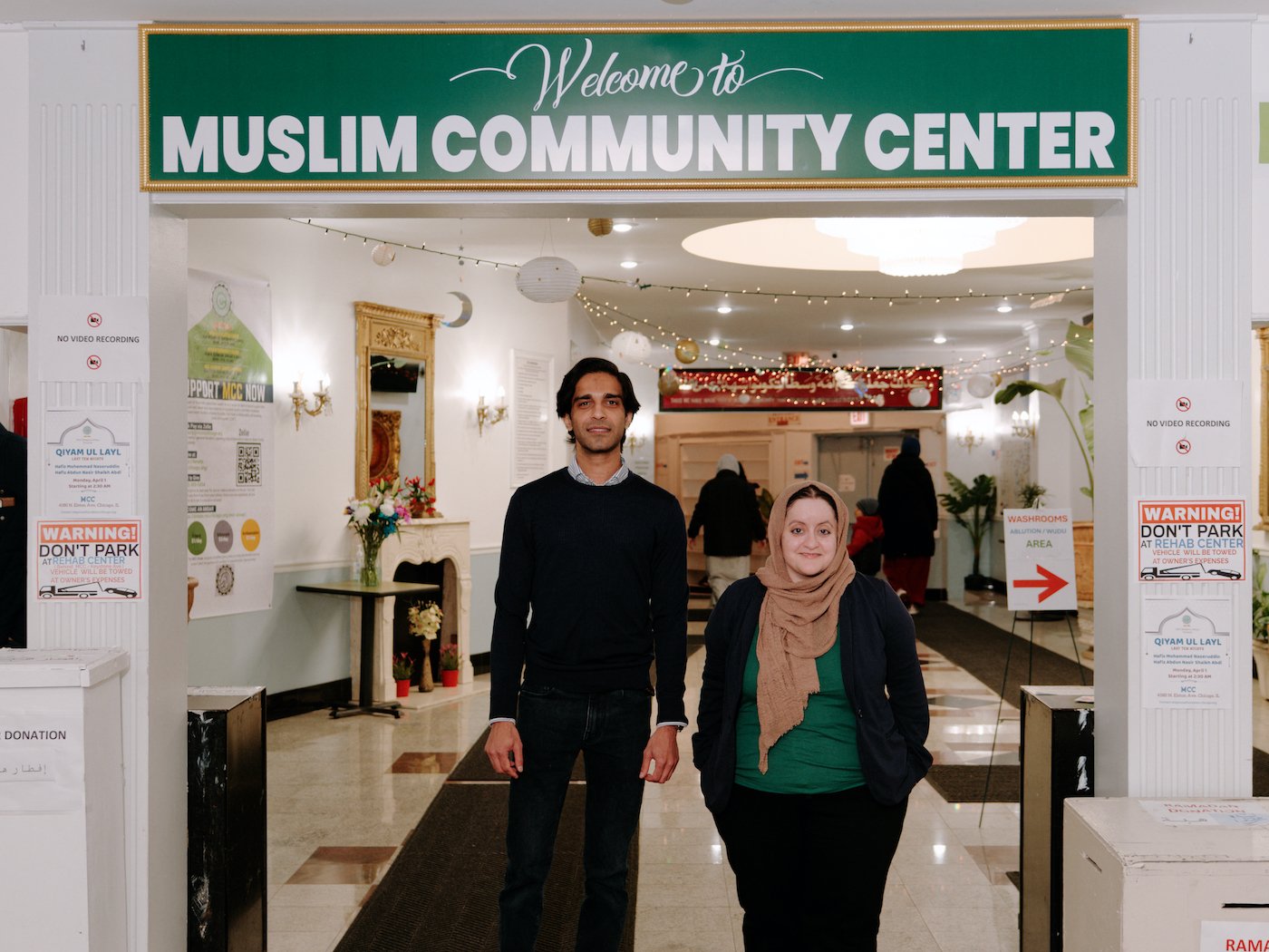Azim Gandhi and Amal Muhsin stand under a sign that says "Welcome to Muslim Community Center"
