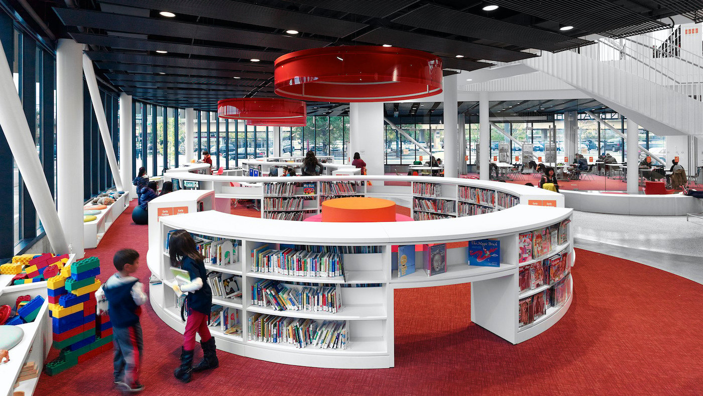 The red-carpeted interior of the Chinatown library, with kids playing with blocks and shelves of books against glass walls