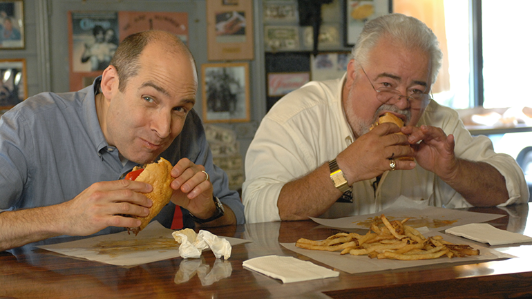 Geoffrey Baer eating Chicago-style hot dog at bar and looking at camera