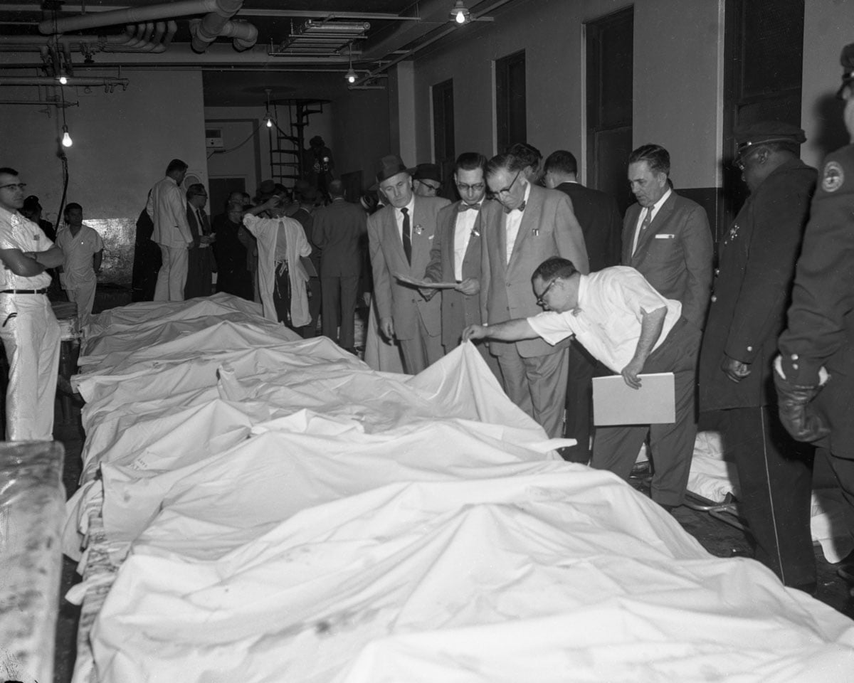 Bodies covered with white sheets lying in a row with officials attempting to identify them