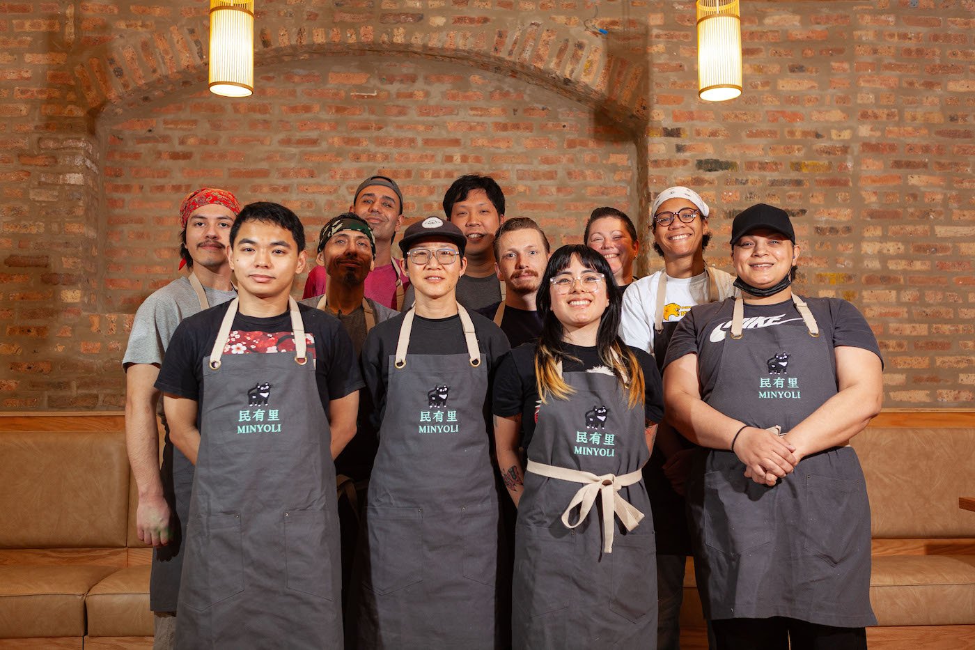 The staff of a restaurant, some in aprons with its name and logo, stand and smile for a group photo