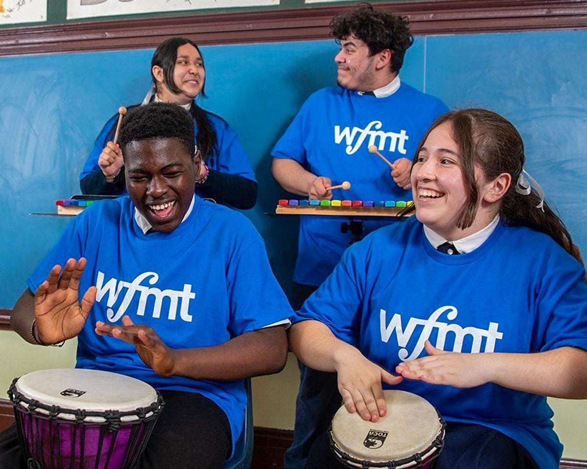 Four students in classroom with WFMT shirts on, happily playing djembe drums and xylophones