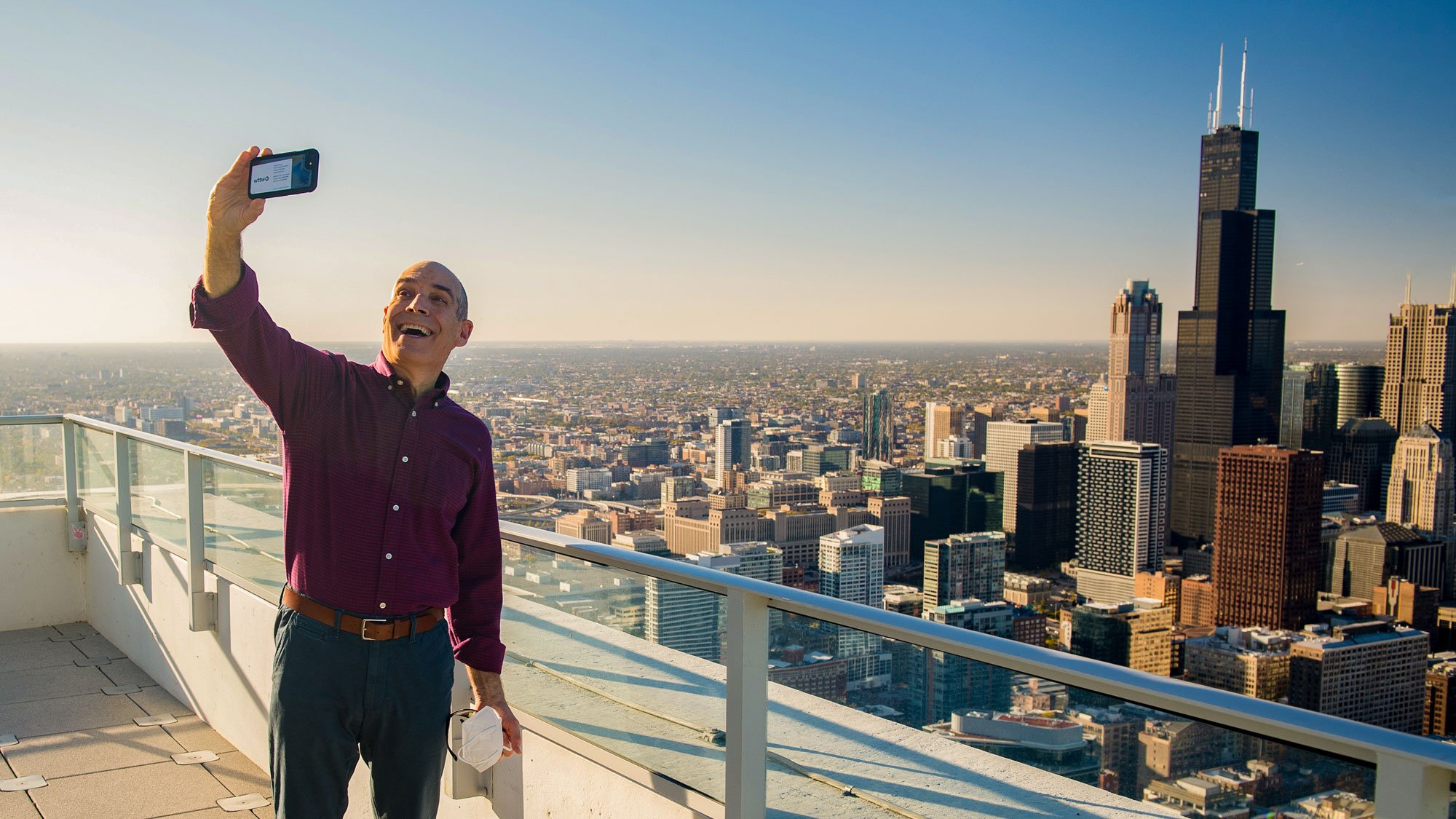 Geoffrey Baer standing on patio of tall building taking selfie with downtown Chicago in background