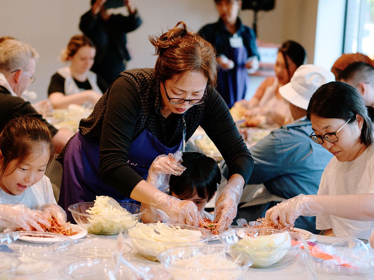 An adult leans over a seated child to help them mix their ingredients on a plate