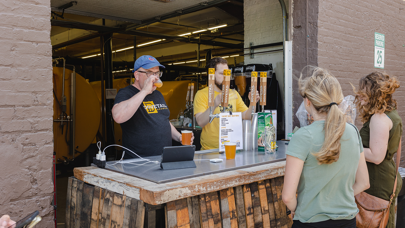 People wait in line to get a beer outside while two men stand behind the bar