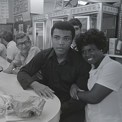 Muhammad Ali seated at a lunch counter, takes picture with fan. Miami, FL. February 25, 1971. Photo: Courtesy of Charles Trainor