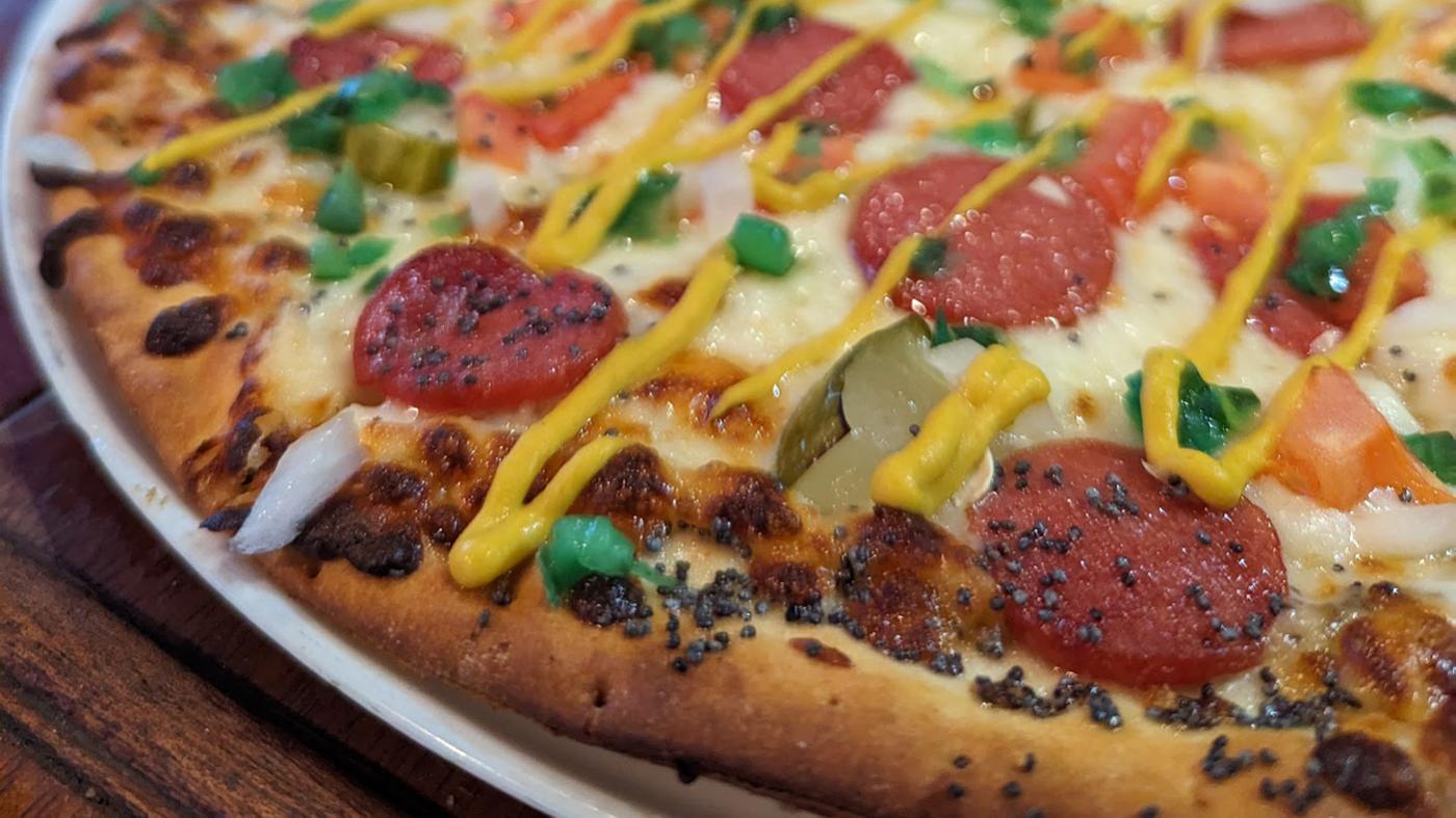 A Chicago Dog Pizza from the Olympic Tavern in Rockford, with a poppy seed and celery salt crust, sliced hot dogs, pickles, tomato, onion, giardiniera, yellow mustard, and relish