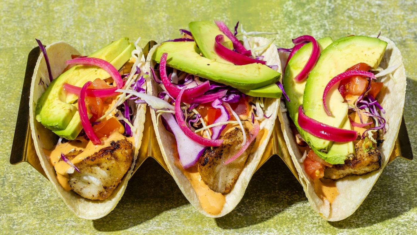 Three fish tacos with avocado and pickled red onion and slaw in a stand