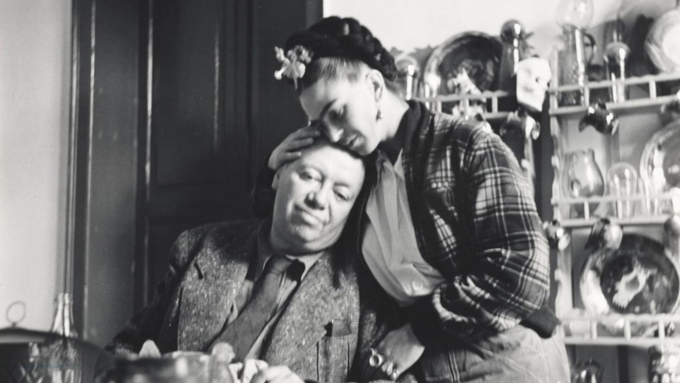 Diego Rivera sits in a kitchen and leans his head against Frida Kahlo, who holds him tenderly