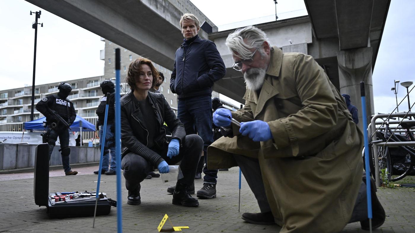Hendrik squats and works on a crime scene while Lucienne squats behind him and Van der Valk stands