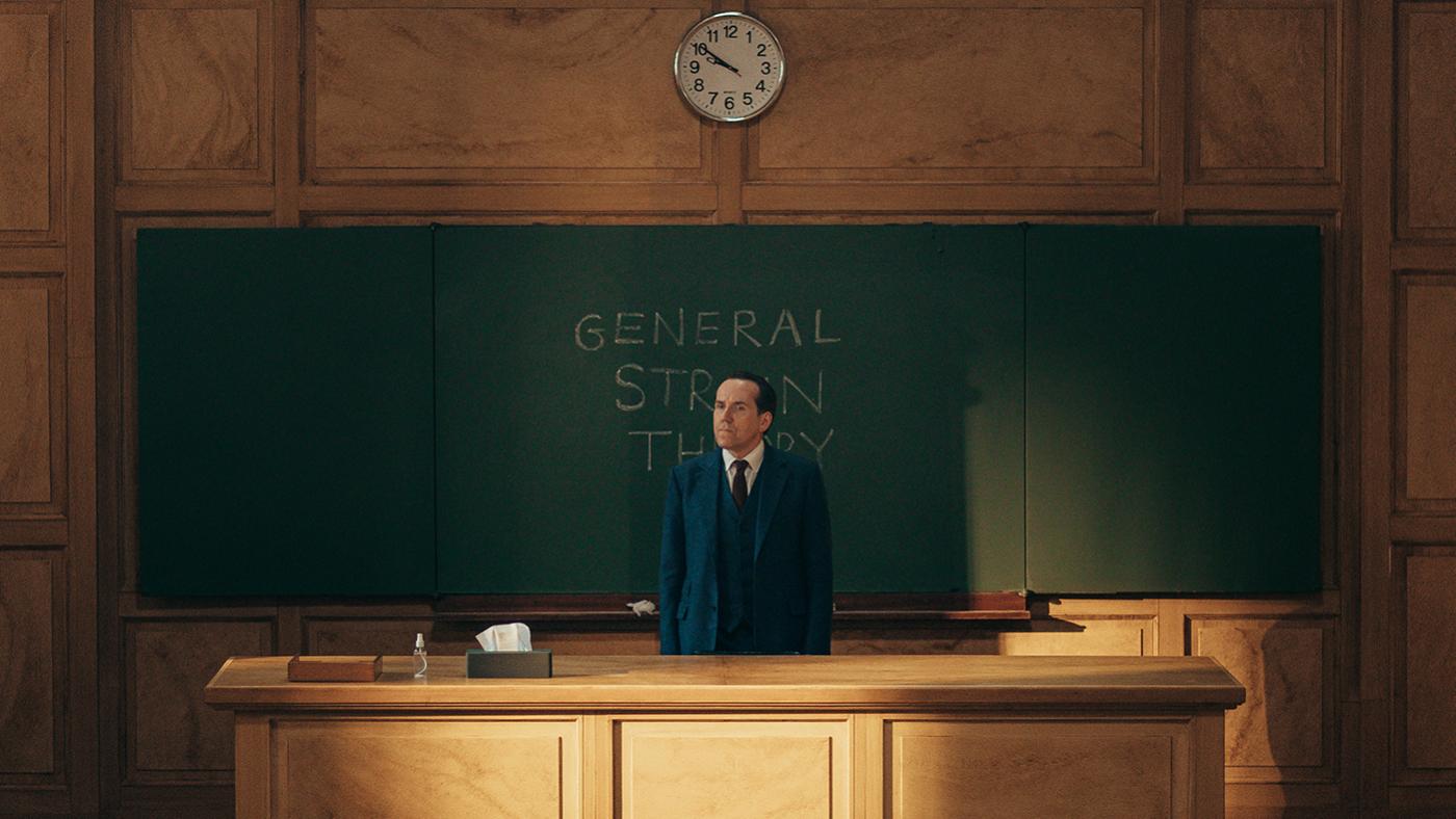 Professor T stands behind a desk in front of a chalkboard in a lecture hall