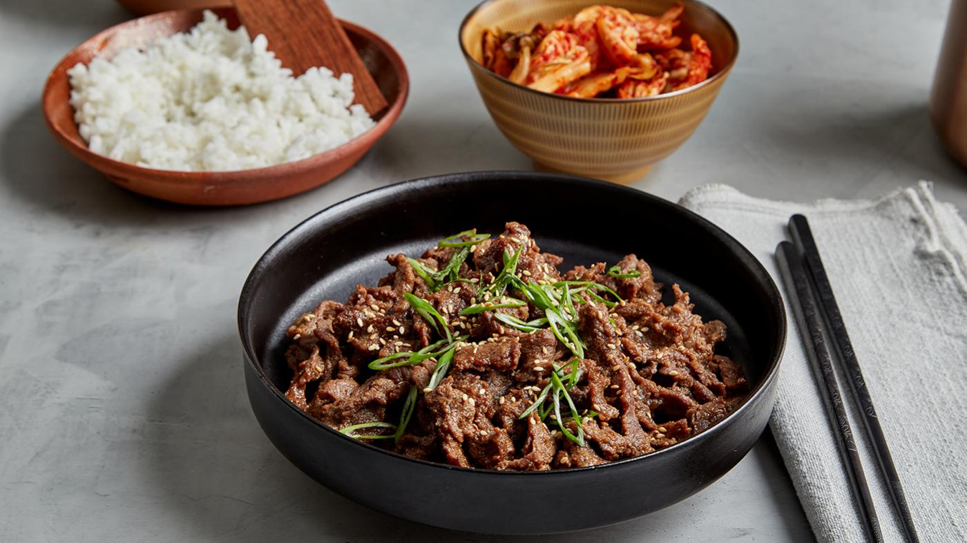 A dish of beef bulgogi on a table with chopsticks on a napkin, kimchi in a bowl, and rice in a dish