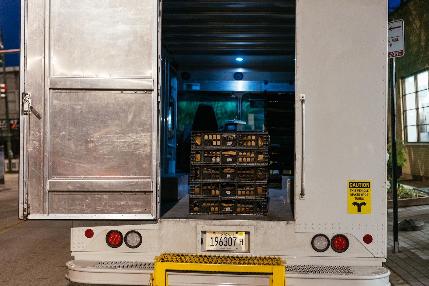 The back door of a delivery truck is open to reveal a stack of pallets containing loaves of bread