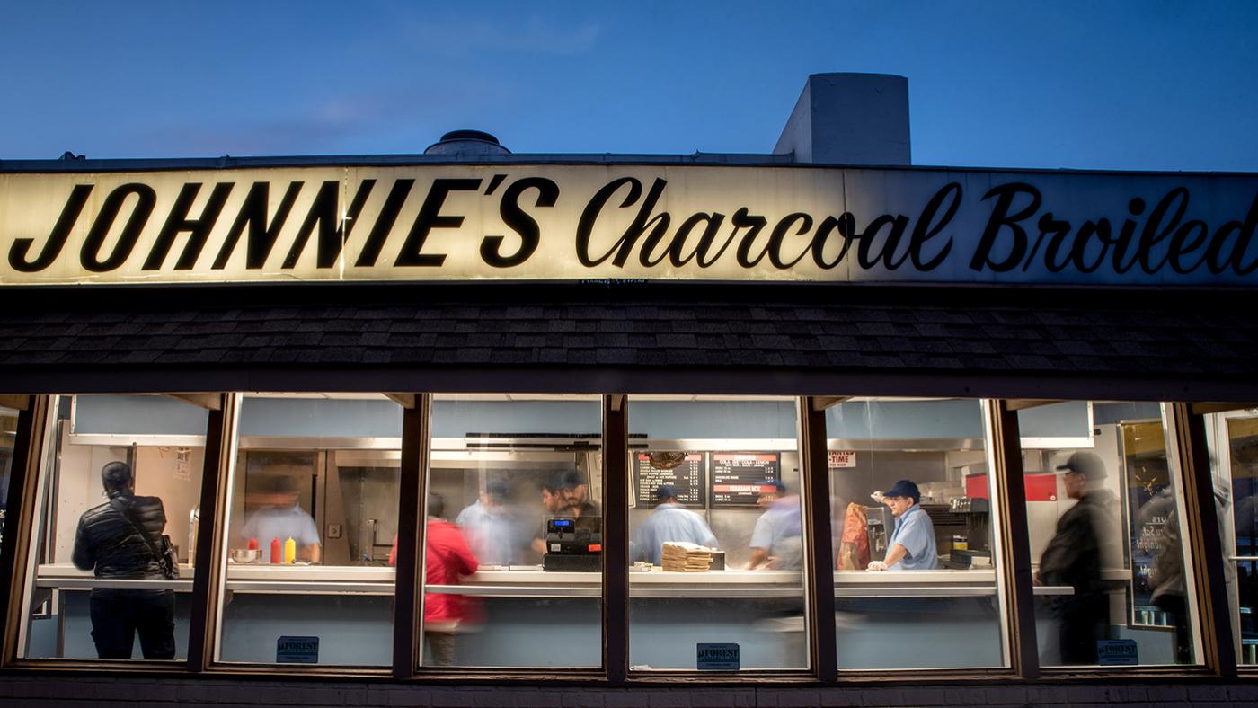 The exterior of Johnnie's beef, looking through the window with blurred figures behind the counter and ordering food. 