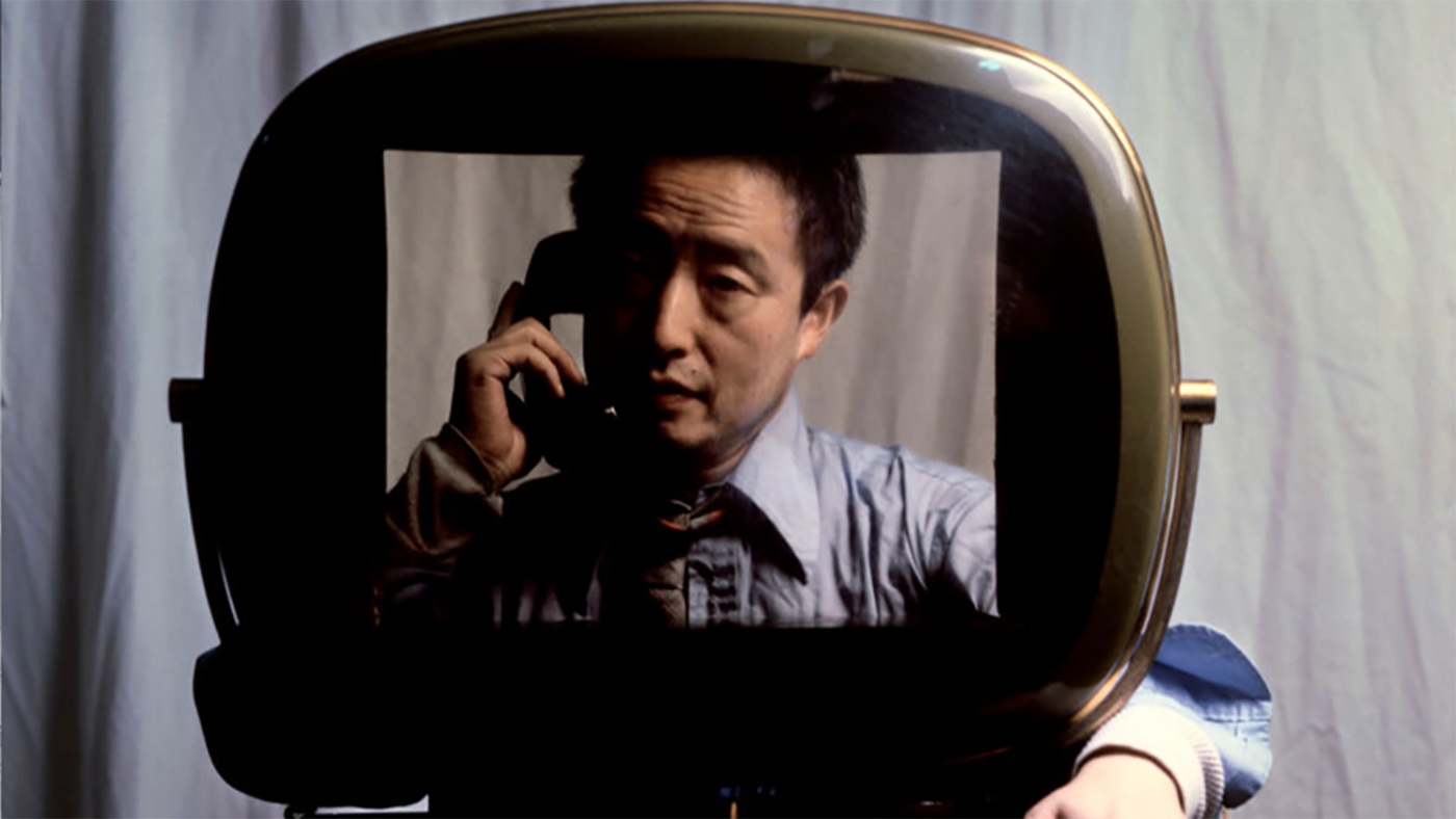 A photo of Nam June Paik sitting behind an open television screen 