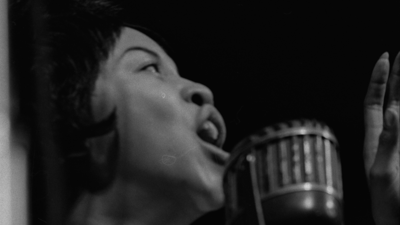 Mavis Staples sings into a microphone with her hand up in a black and white 1962 photograph