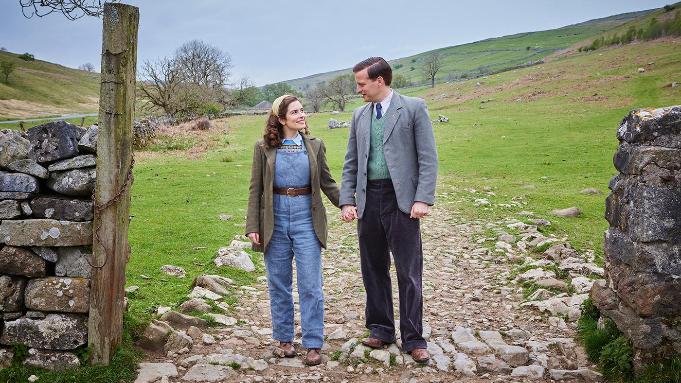 Helen and James Herriot happily look at each other as they stand on a path between two stone walls