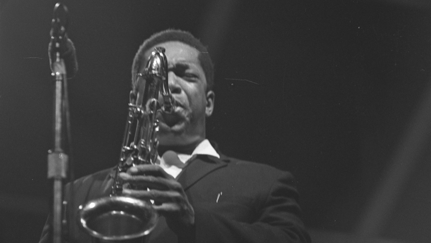 John Coltrane plays saxophone in front of a mic with a black background in 1965