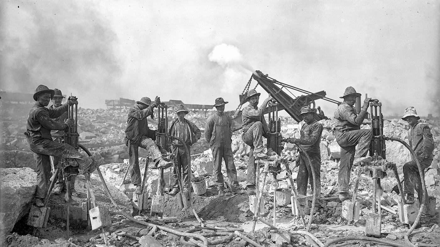 A group of men stand with heavy equipment amongst rock in a black and white photo from 1904