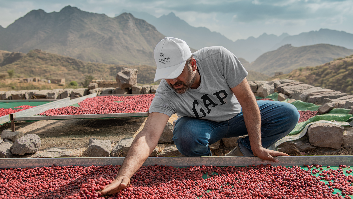 A man squats next to a tray of coffee fruit drying in the sun in front of mountains
