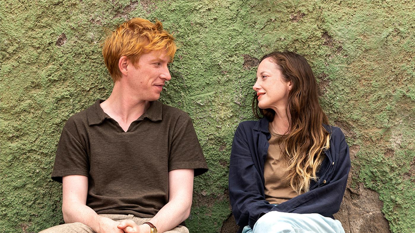 Jack and Alice look at each other while sitting on a bench against a green wall