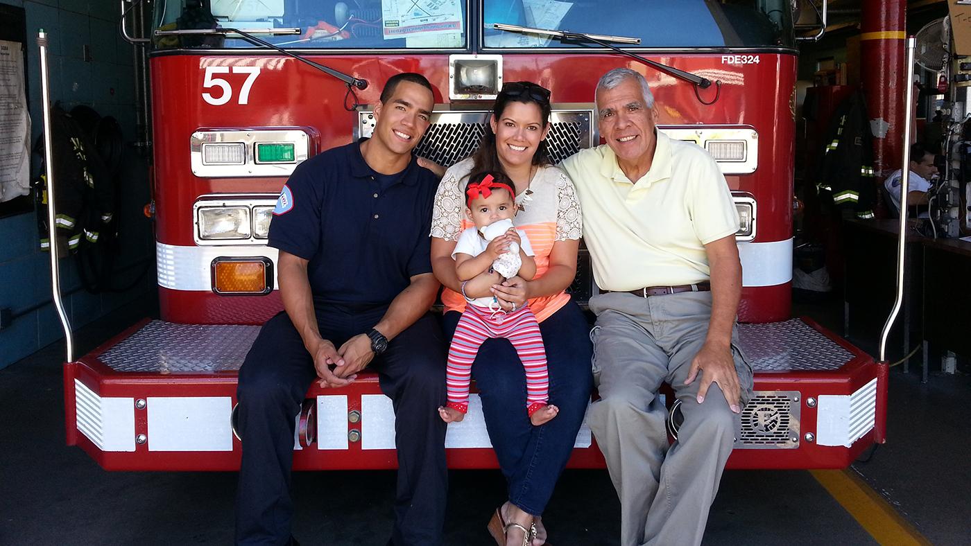 Elan, Bianca, and Carlos Lozano sitting on the front of a fire truck in 2014. Bianca is holding her daughter, Anabella. 