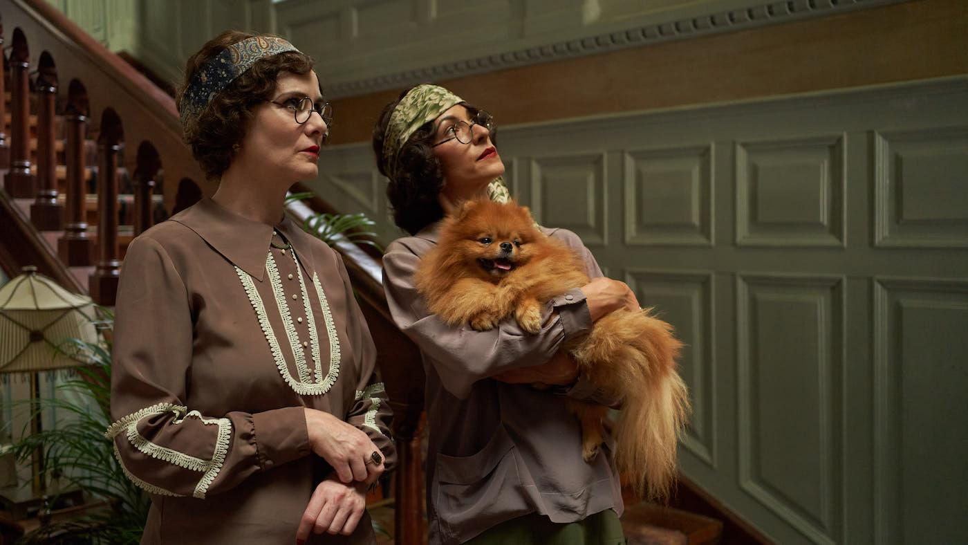 Two women in flapper dress and glasses look up while one holds a Pomeranian dog