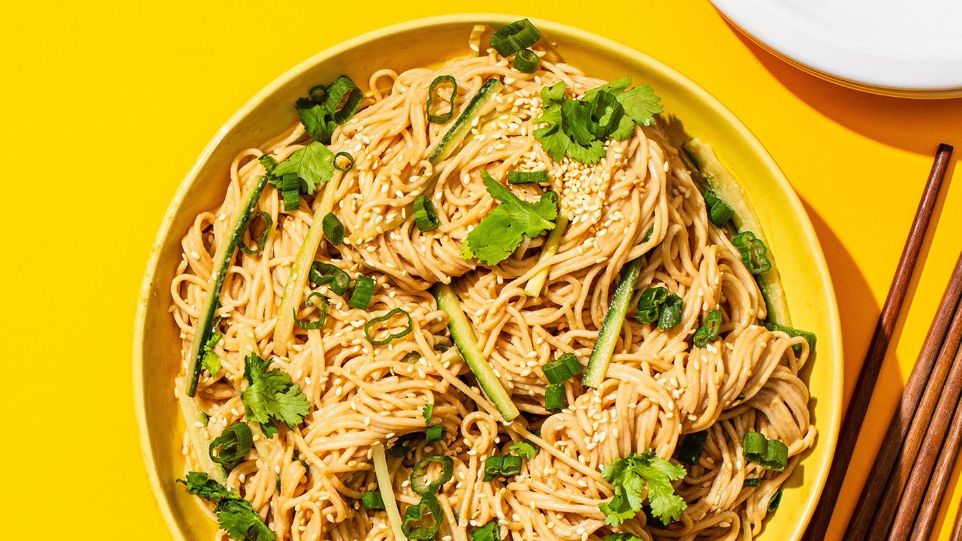 A bowl of cold sesame noodles topped with cilantro, scallions, and cucumber in a yellow bowl on a yellow background