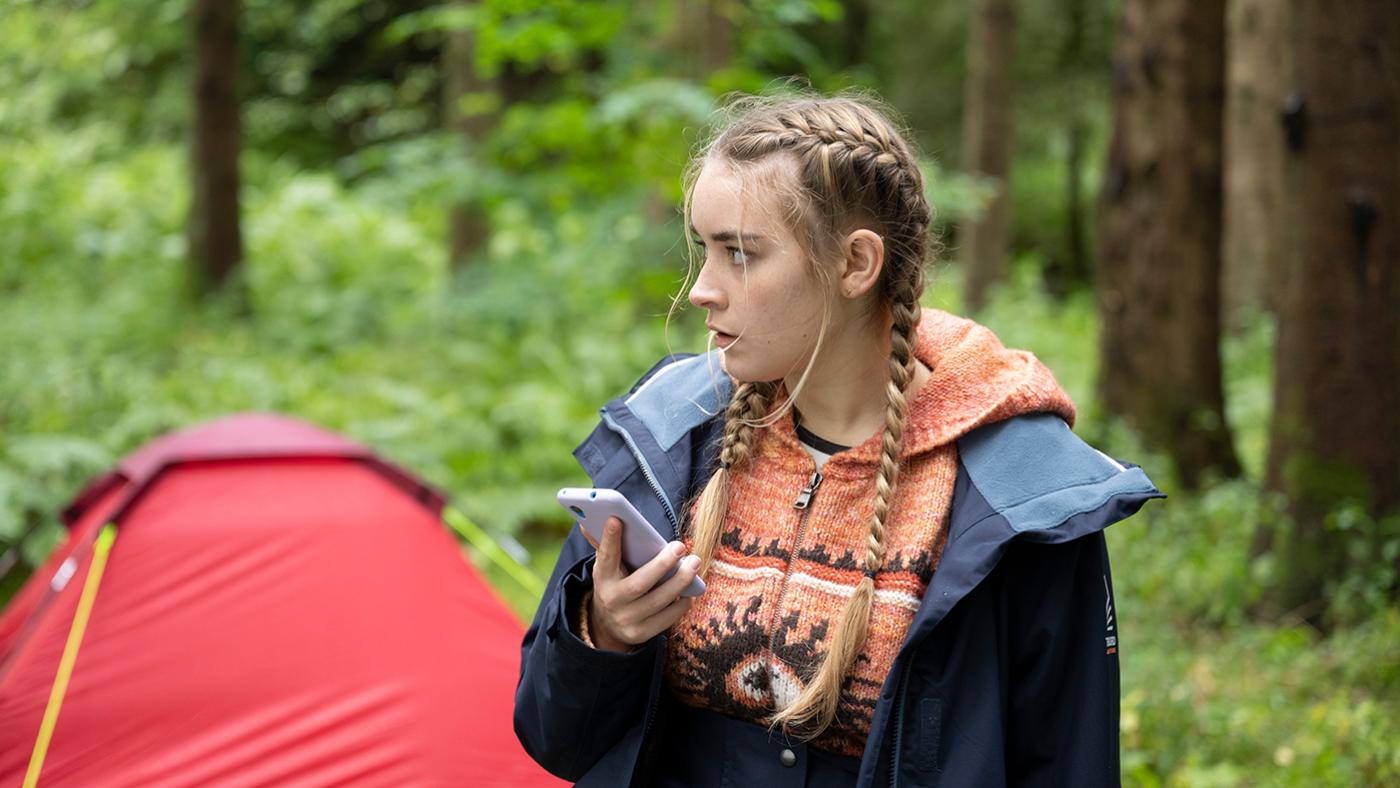 Morgan holds her phone and stands in front of a tent in a forest