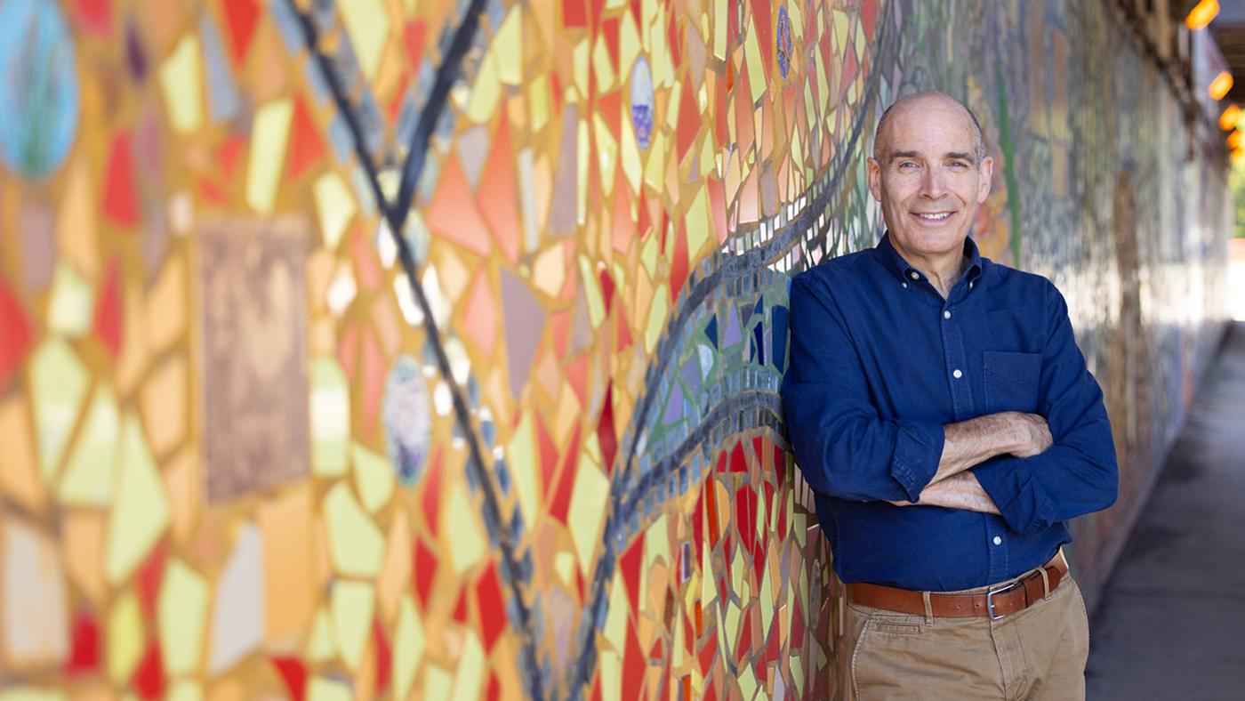 Geoffrey Baer stands in front of a colorful mosaic mural