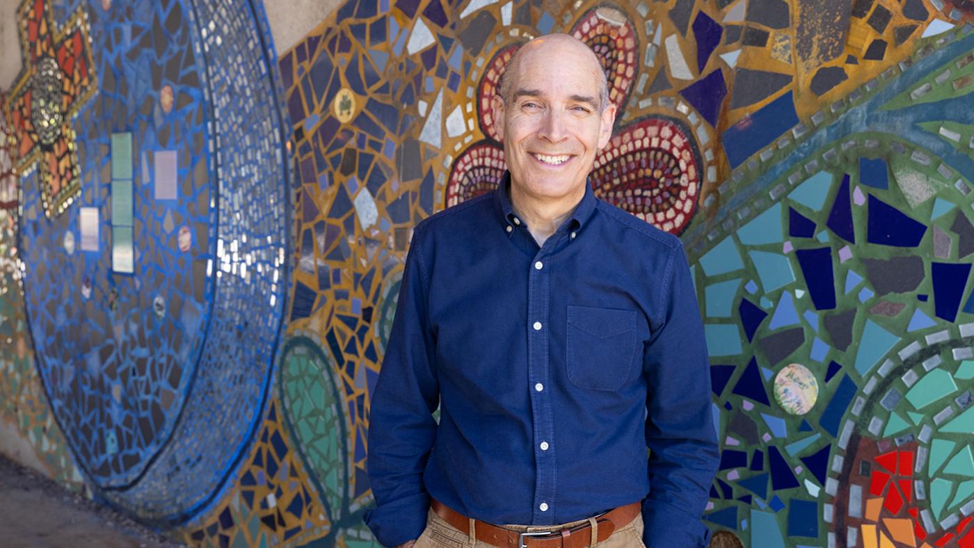 Geoffrey Baer smiles in front of a colorful mosaic mural