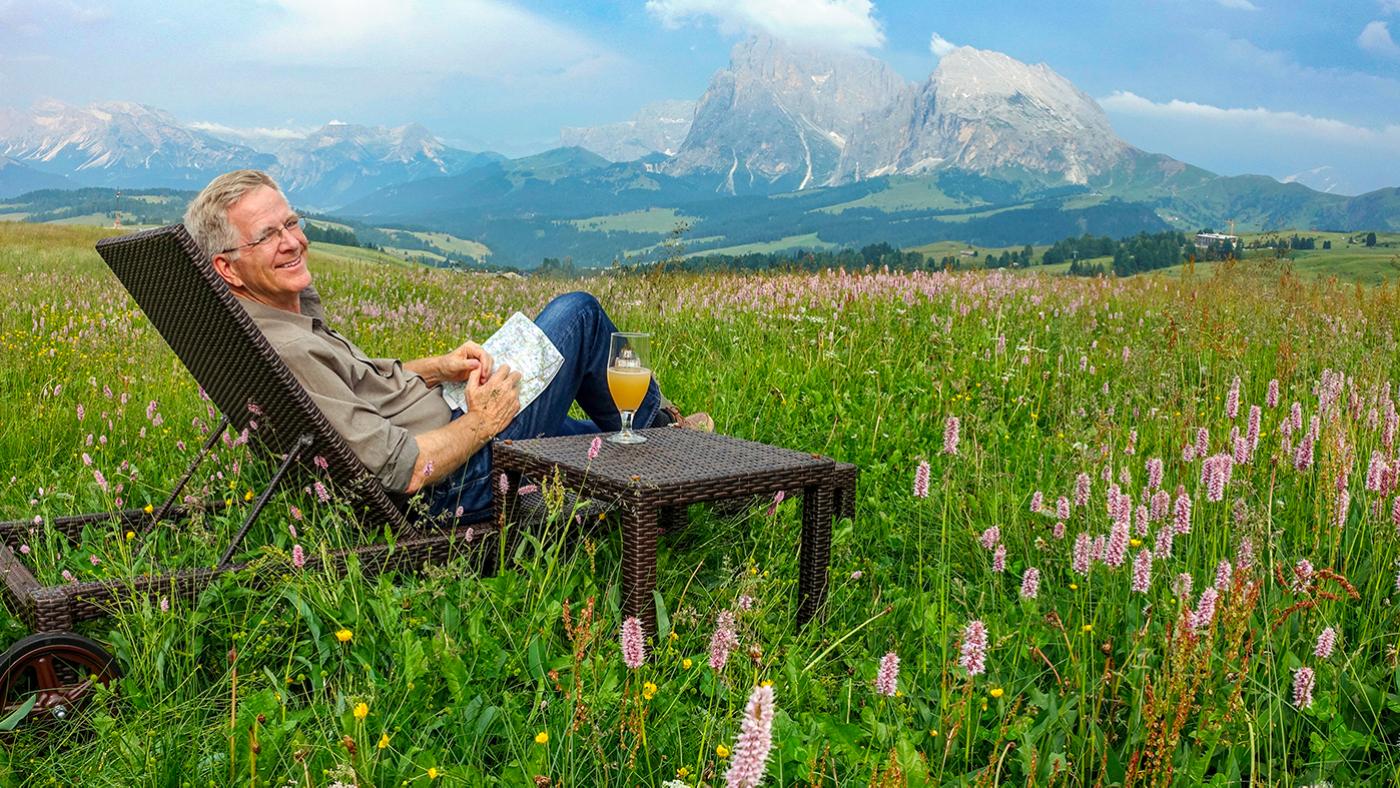 Rick Steves relaxes with a beer in a field in front of a mountain