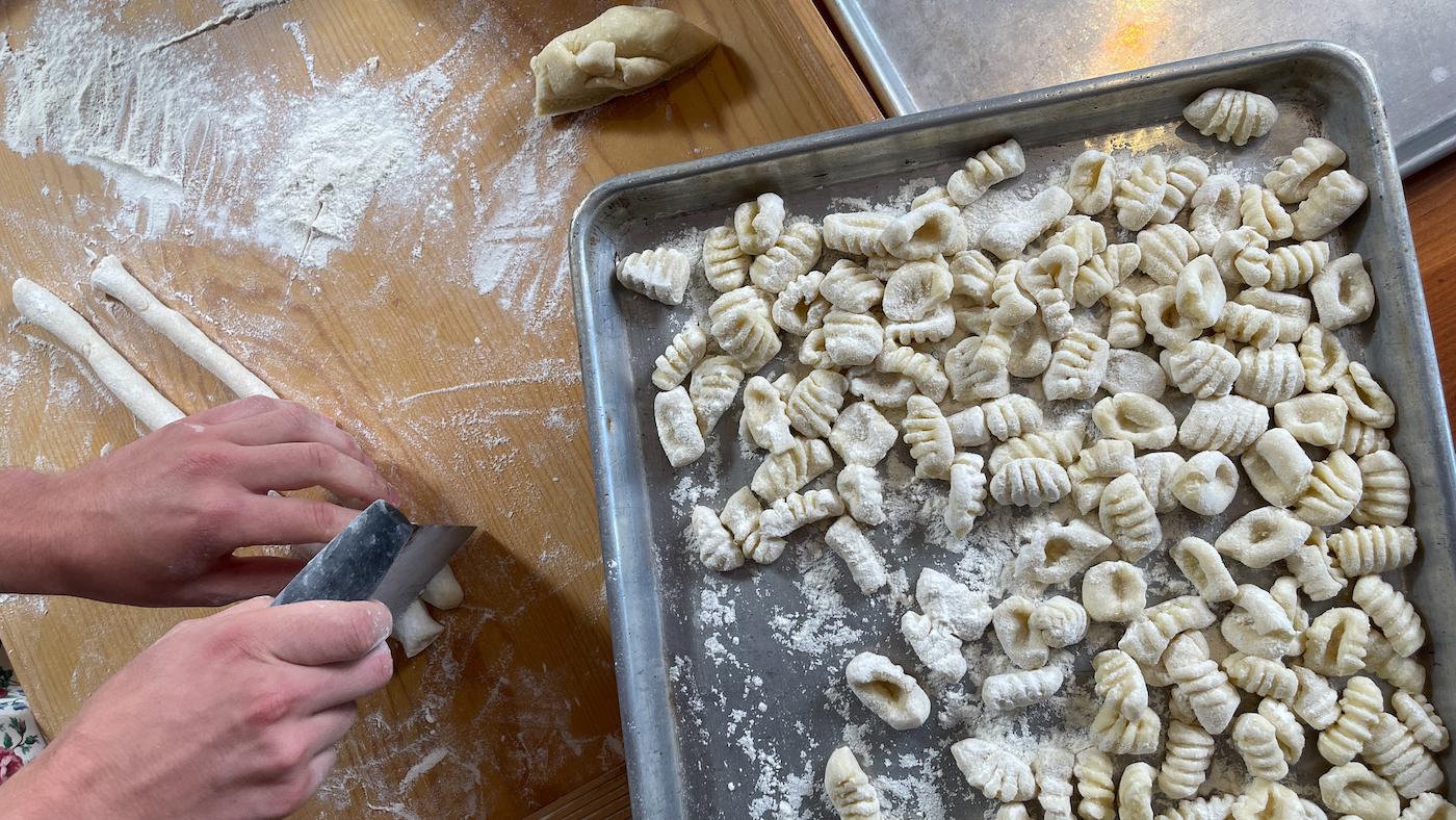 Uncooked potato gnocchi sit on a tray while hands cut more from dough