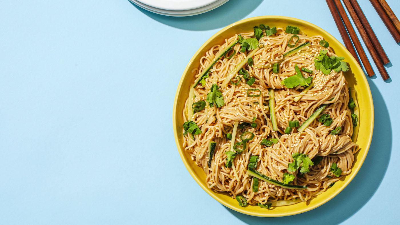A yellow bowl of sesame noodles on a blue background with chopsticks