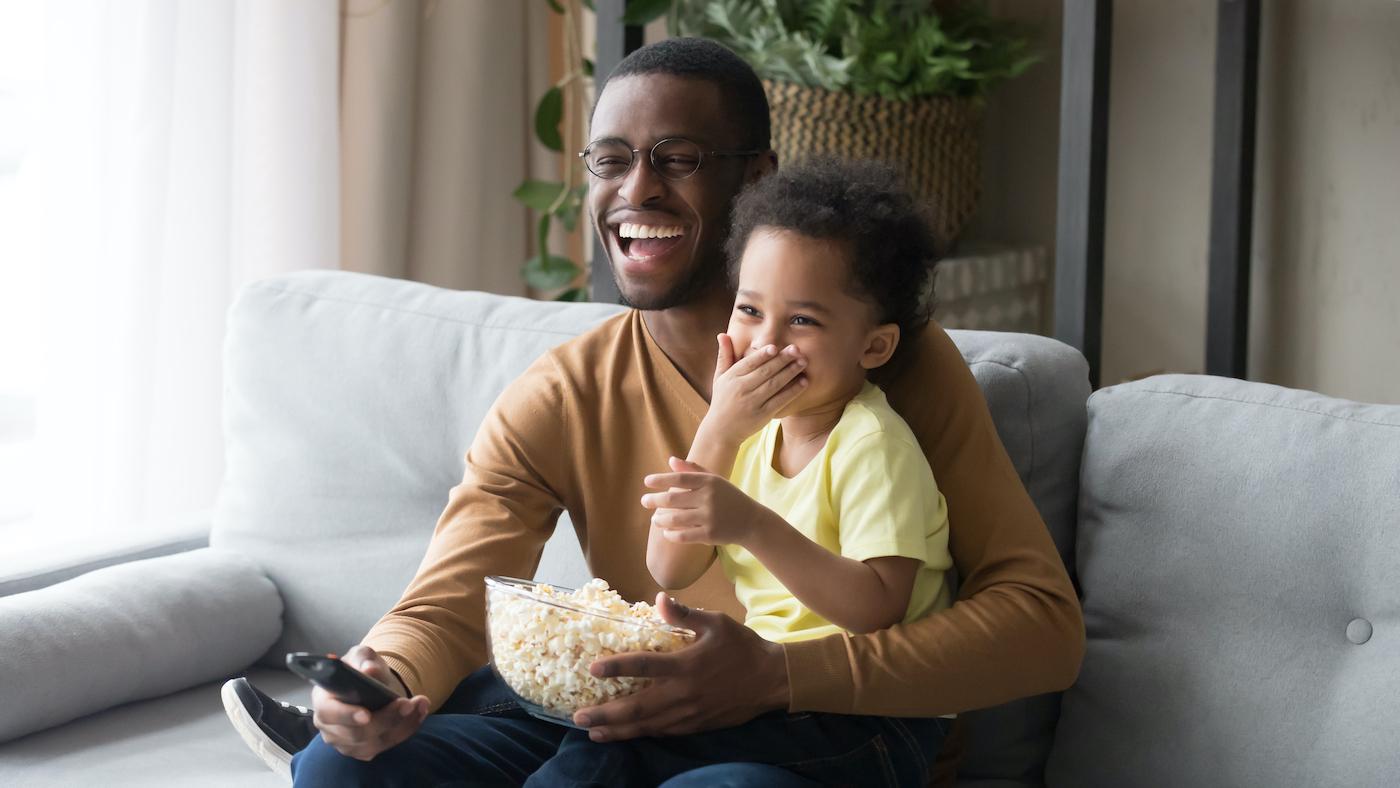 A Black father and son enjoy a bowl of popcorn on the couch and watch TV