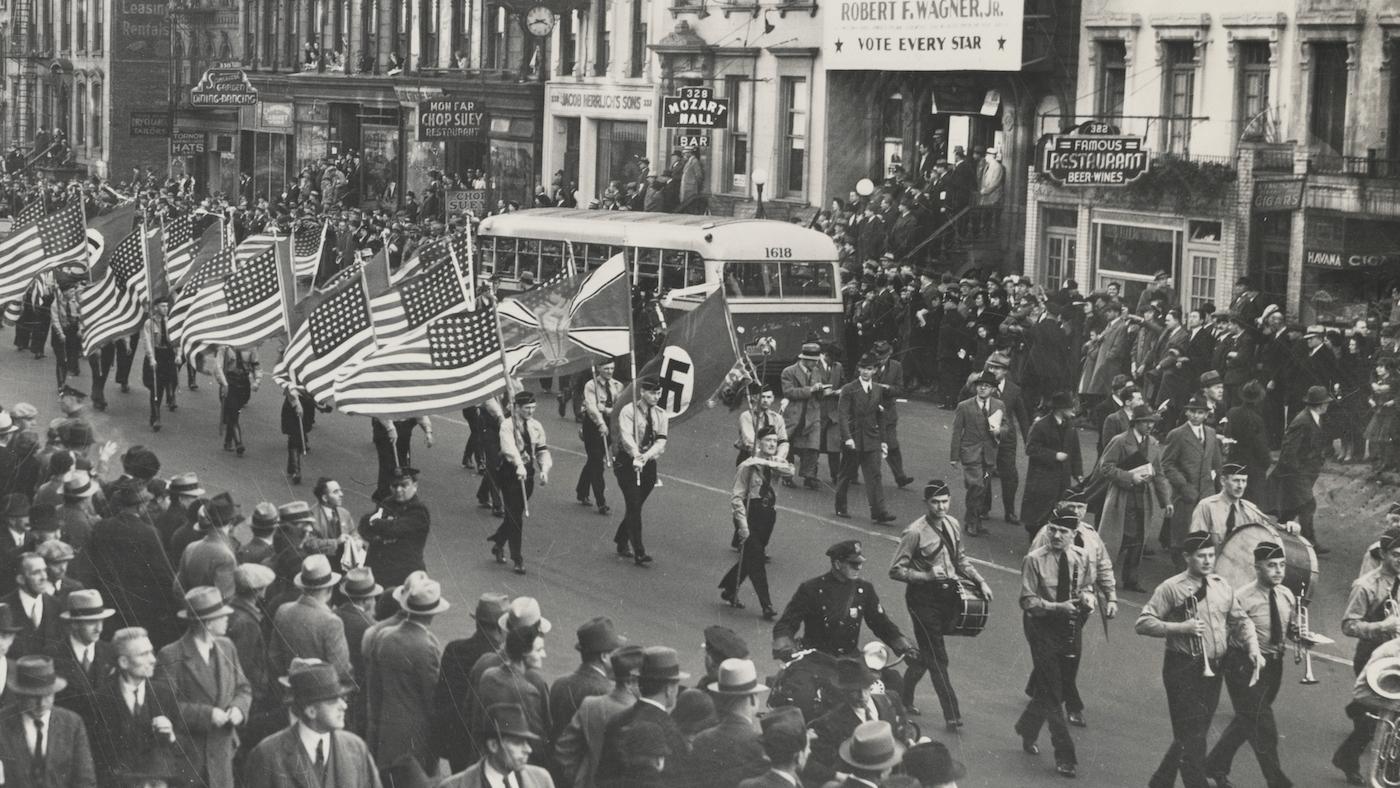 A parade down a crowded Manhattan street in the 1930s featuring American flags and a swastika in a black and white photo