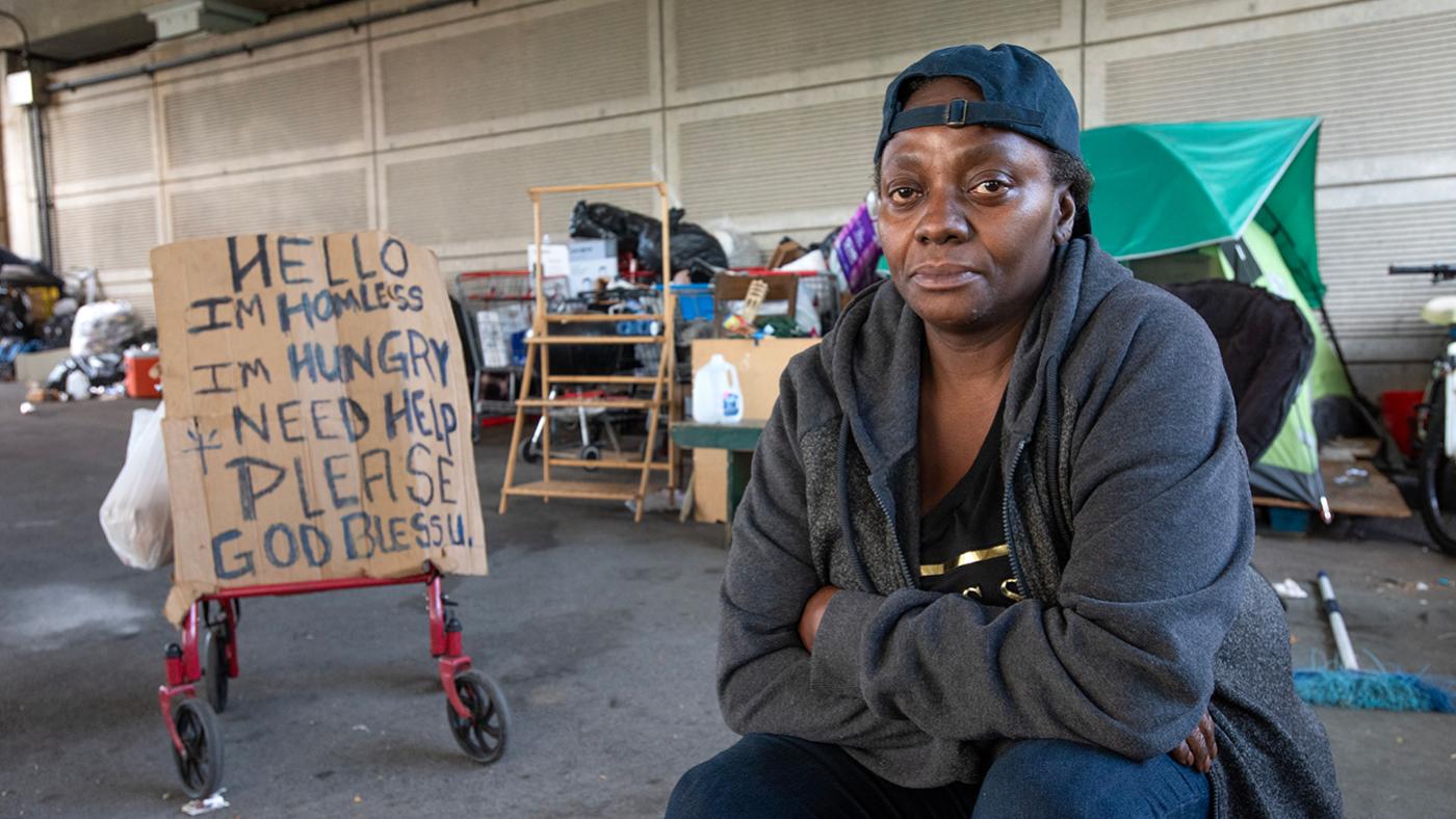 Yolanda sits near her cardboard sign on a walker asking for help in front of an encampment of people experiencing homelessness