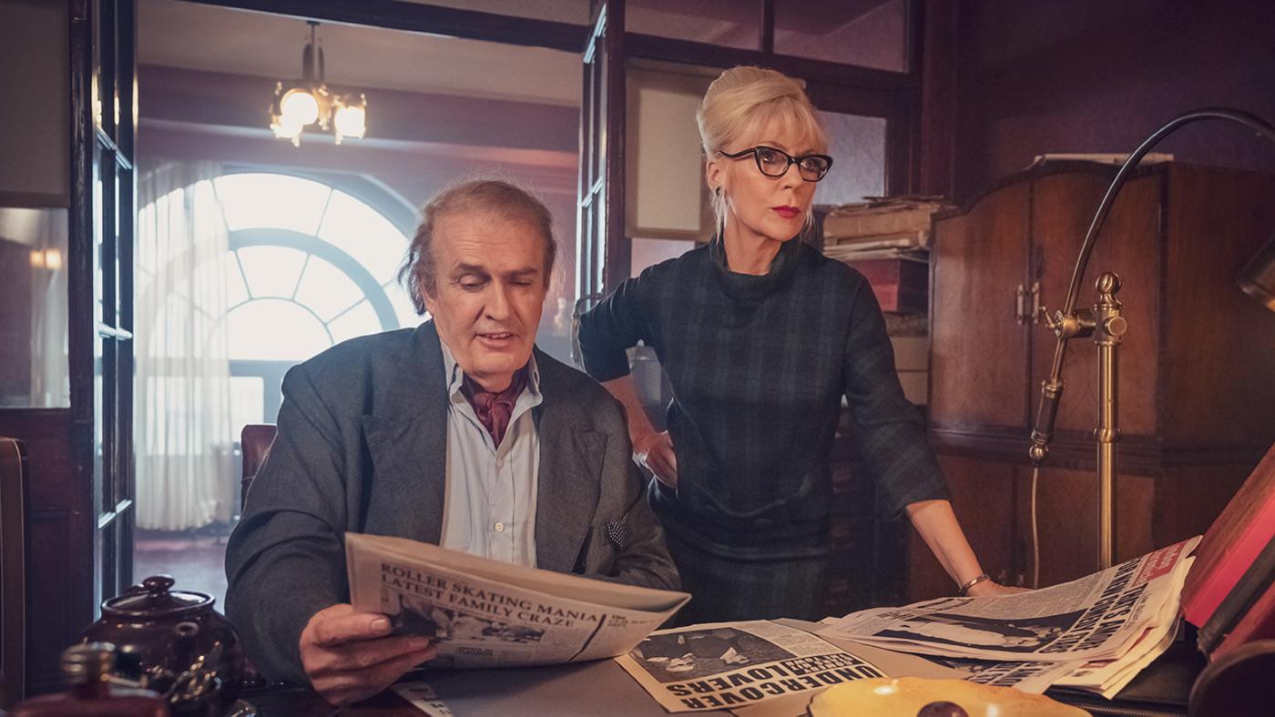 Brian sits at a desk looking at a newspaper while Patsy stands next to him