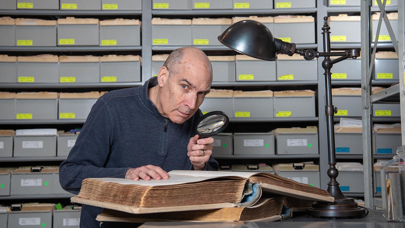 Geoffrey Baer looks at an old record book under a lamp with a magnifying glass