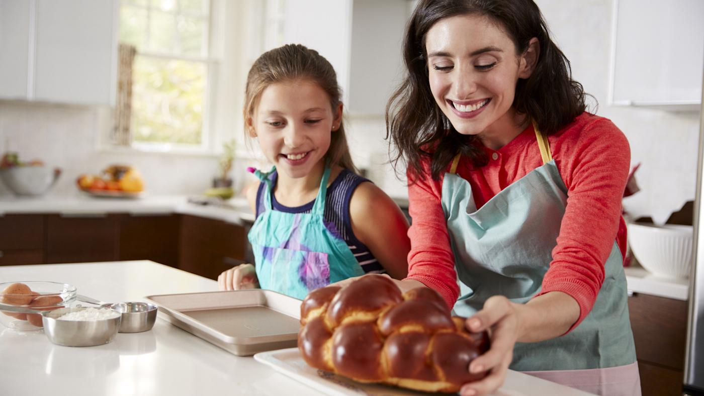 A mother and daughter in aprons smile at a freshly baked loaf of challah